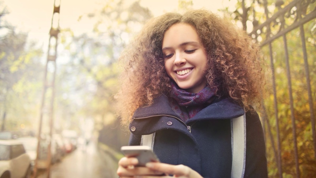 A young woman smiling as she reads a message on her phone outdoors in warm autumn light